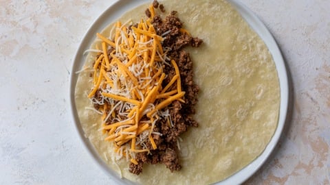 A flour tortilla on a white plate is topped with cooked ground beef and a generous amount of shredded yellow and white cheese, ready to be folded into a quesadilla.