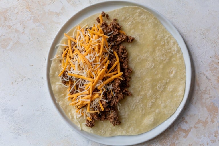 A flour tortilla on a white plate is topped with cooked ground beef and a generous amount of shredded yellow and white cheese, ready to be folded into a quesadilla.