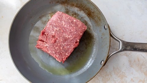 Raw ground beef is placed in a lightly oiled frying pan, ready to be cooked. The pan sits on a light-colored surface.