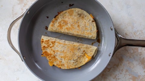 Two folded quesadillas cooking in a gray nonstick skillet. The quesadilla is golden brown with some cheese melting out of the edges. The pan is on a light textured surface.