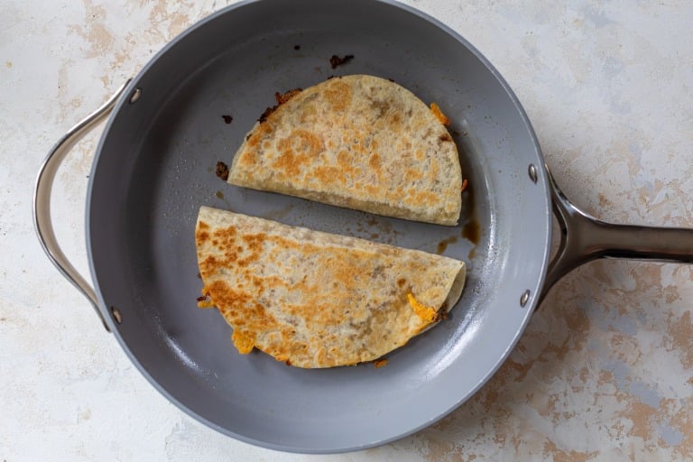 Two folded quesadillas cooking in a gray nonstick skillet. The quesadilla is golden brown with some cheese melting out of the edges. The pan is on a light textured surface.
