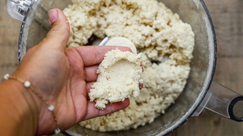 A hand holding a pinch of crumbly dough over a food processor filled with more dough.