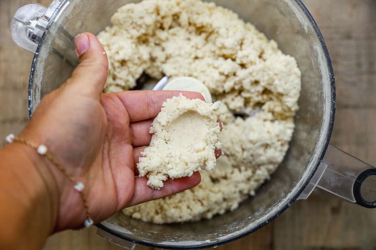 A hand holding a pinch of crumbly dough over a food processor filled with more dough.