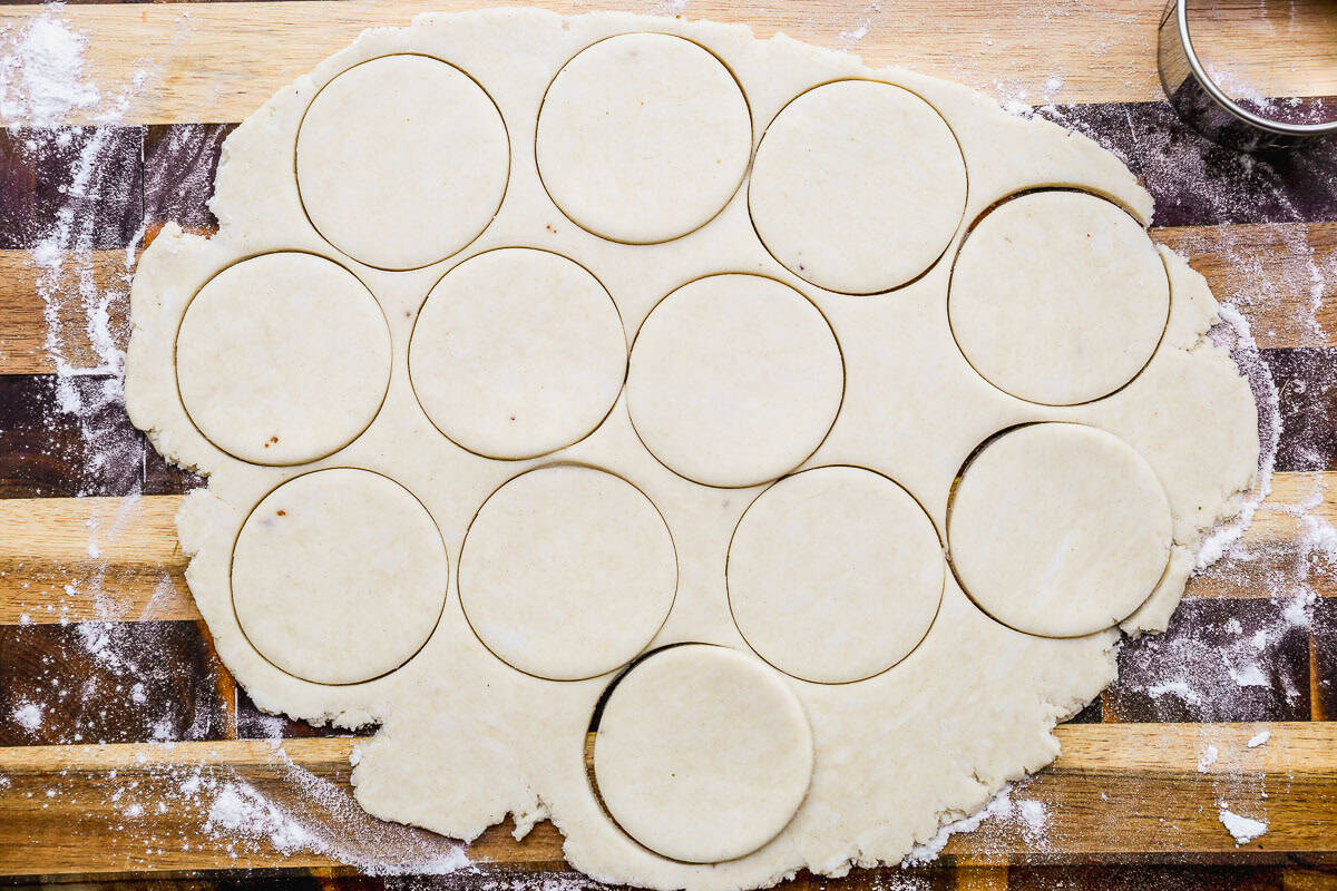 Rolled-out dough with round shapes cut out, resting on a floured wooden surface with a metal cutter nearby.