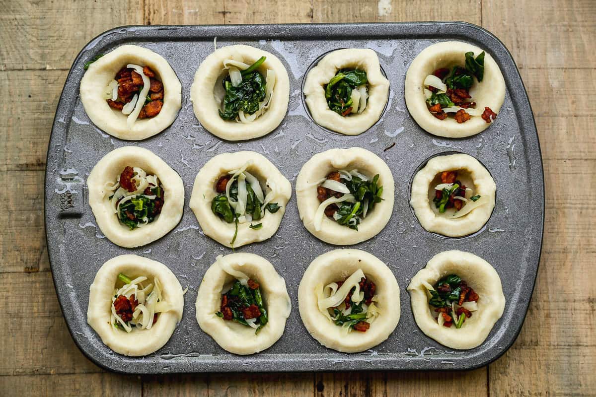A muffin tin filled with dough cups, some with cheese, spinach, and bacon on a wooden surface.