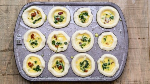 A muffin tin filled with unbaked mini quiches, containing egg mixture, spinach, and sun-dried tomatoes, on a wooden surface.