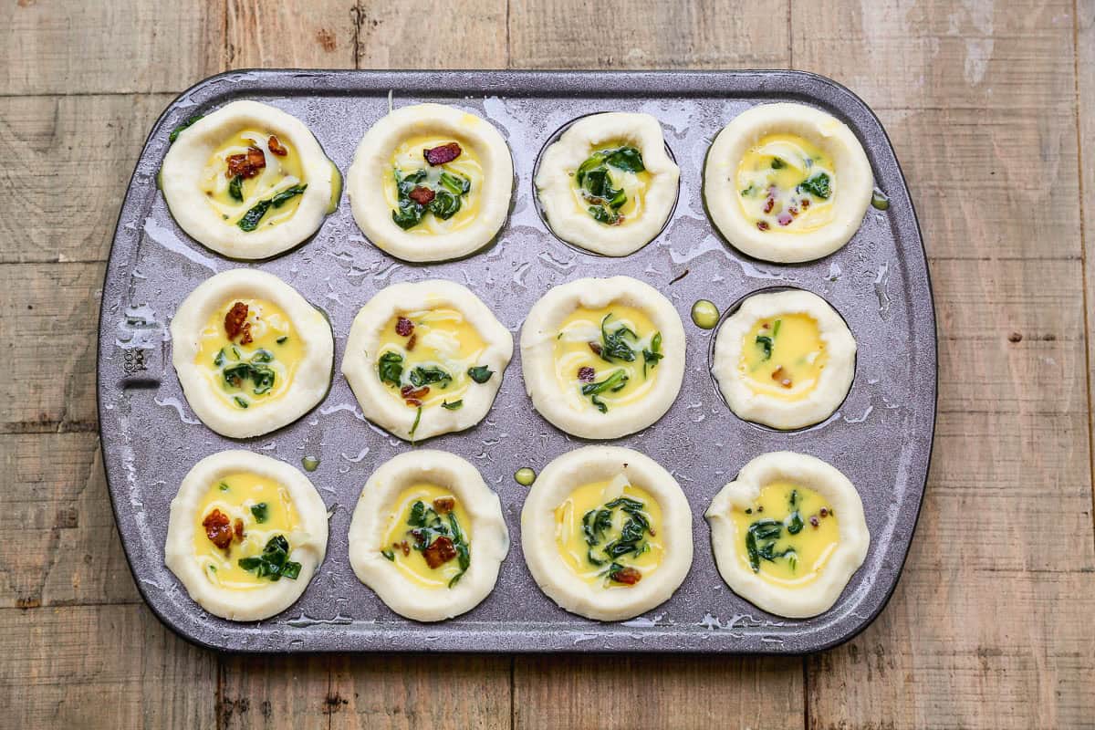 A muffin tin filled with unbaked mini quiches, containing egg mixture, spinach, and sun-dried tomatoes, on a wooden surface.