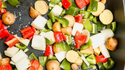 Chopped red and green bell peppers, white onions, and small potato pieces are spread on a dark surface, possibly a pan, ready for cooking.