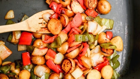 A wooden spatula stirs sautéed vegetables, including halved baby potatoes, red and green bell peppers, and onions, in a dark pan. The vegetables appear lightly browned and cooked.