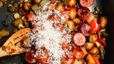 A skillet filled with sautéed sausage slices, diced potatoes, peppers, onions, and mushrooms, topped with a generous sprinkle of grated cheese. A wooden spatula is visible on the left side of the pan.