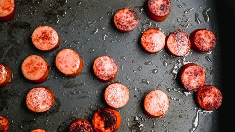 Round slices of sausage being browned in a skillet, with some pieces showing a seared, crispy surface and glistening with oil.
