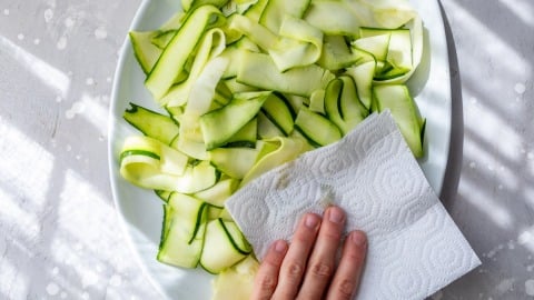 A hand patting zucchini ribbons dry with a paper towel on a white plate.