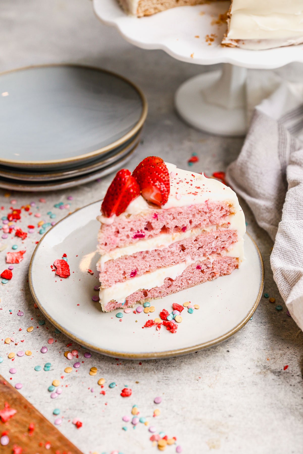 A slice of pink cake with white frosting and fresh strawberries on top, served on a white plate with colorful sprinkles scattered around on the table.