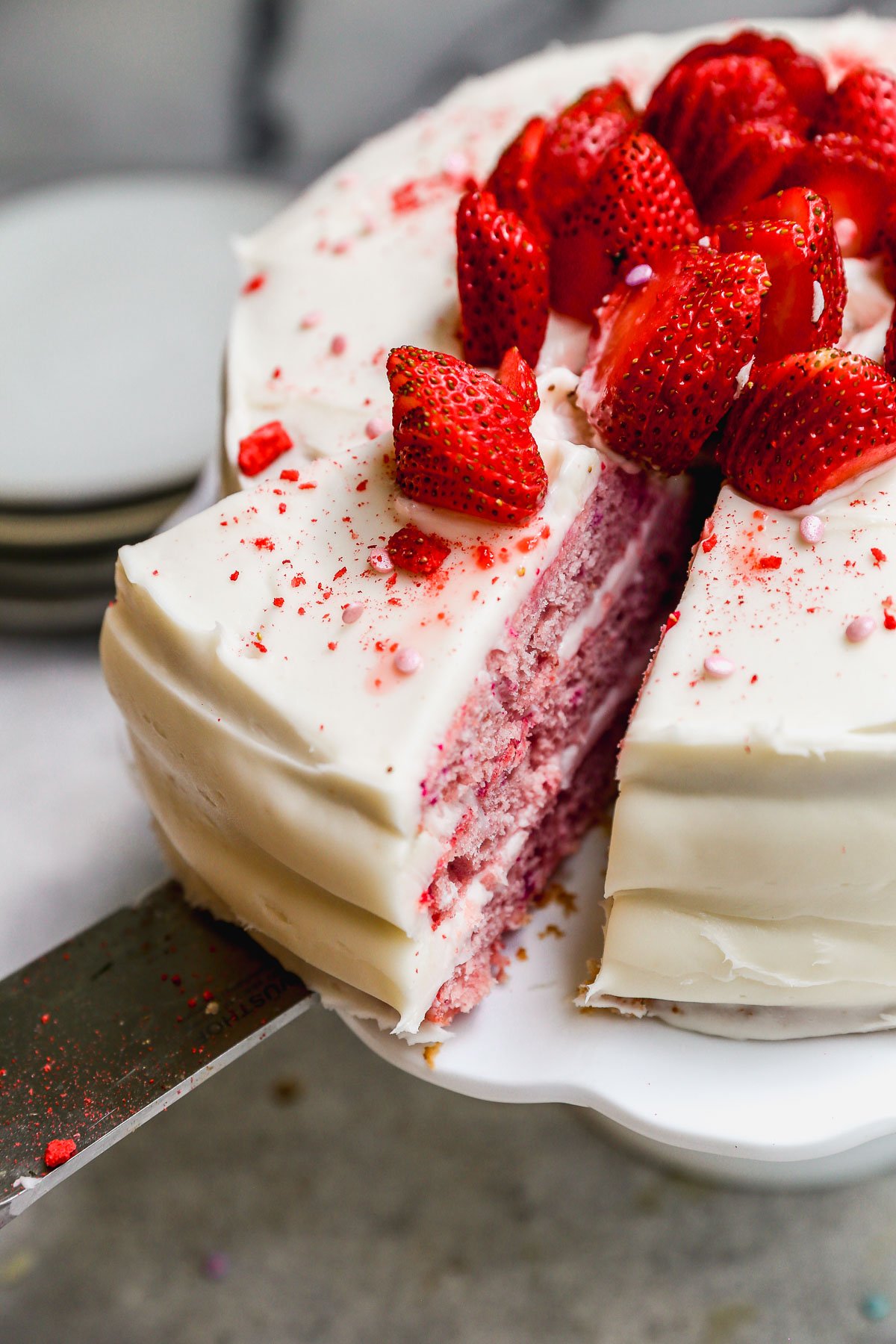 A layered pink cake with white frosting, topped with fresh strawberries, sits on a stand. A slice is being lifted out, revealing pink cake layers inside.