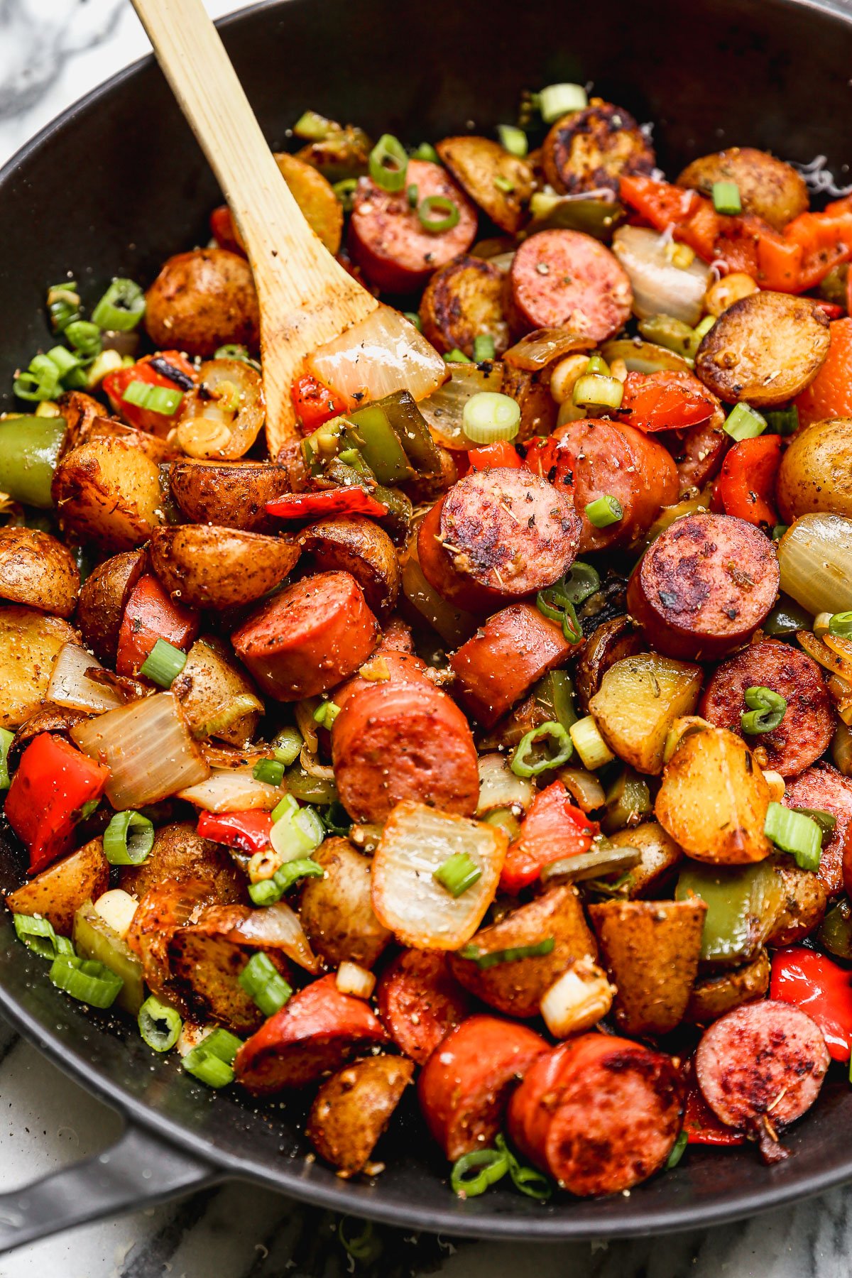 A skillet filled with fried sausage, potatoes, bell peppers, and onions, garnished with chopped green onions, with a wooden spoon stirring the mixture.