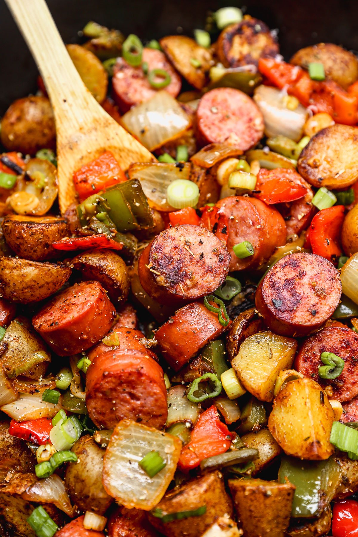 A close-up of a skillet filled with sliced sausage, potatoes, bell peppers, and onions, topped with chopped green onions and stirred with a wooden spoon.