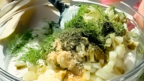 A hand mixes chopped pickles, fresh dill, and spices into a bowl of creamy white sauce with a spatula.