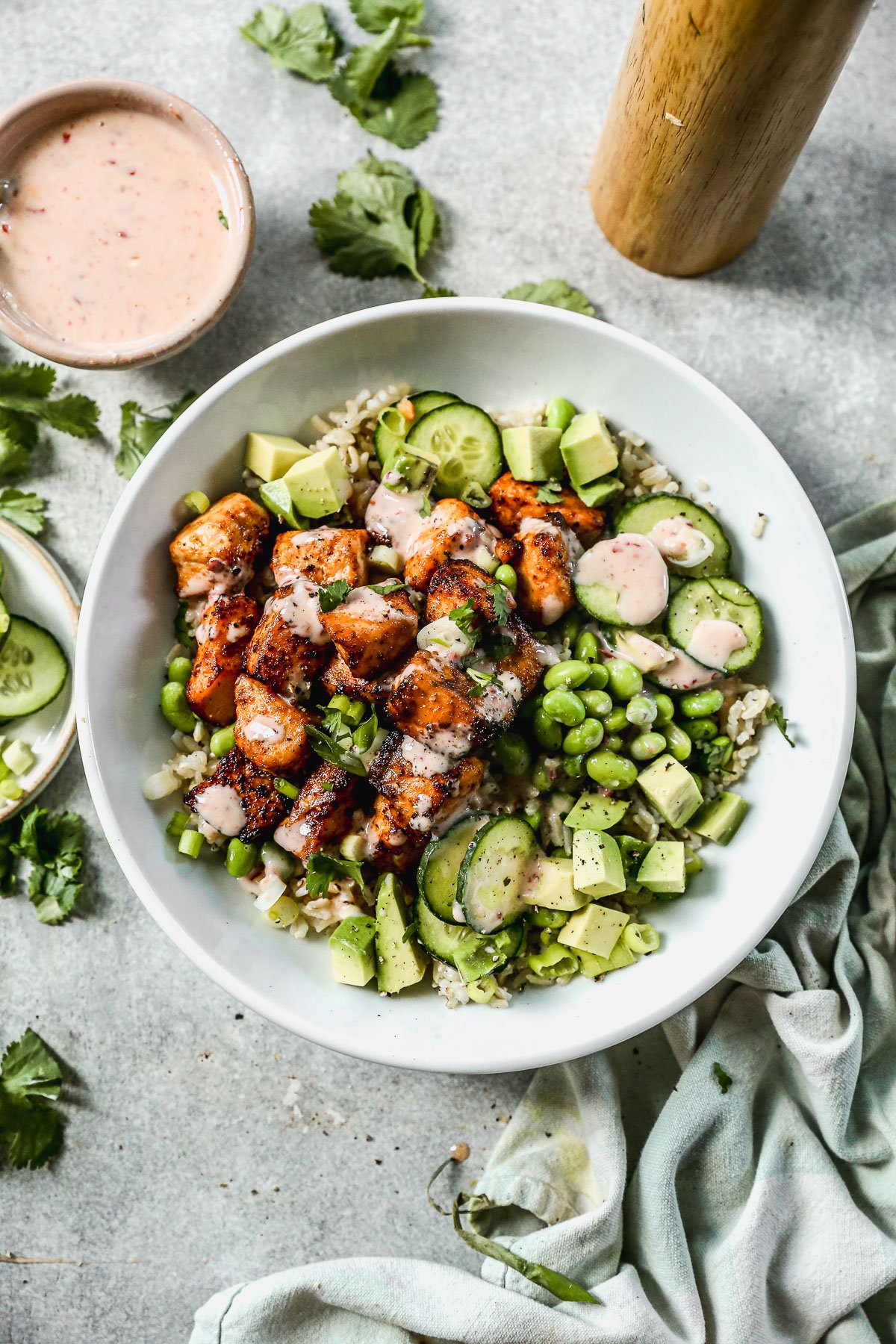 A bowl of bang bang salmon, rice, avocado, cucumber, edamame, and sauce, garnished with cilantro.