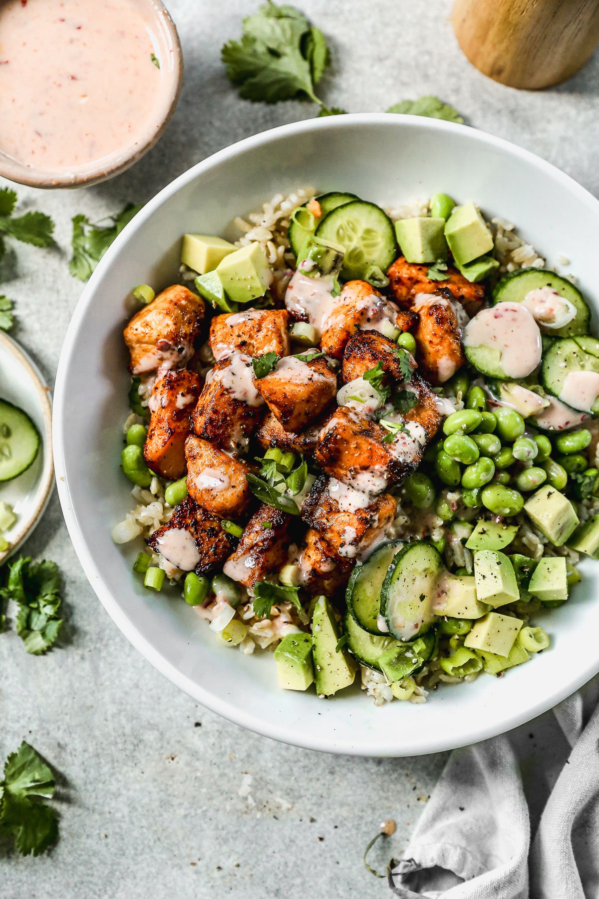 A bowl with bang bang salmon, sliced cucumber, avocado, edamame, rice, cilantro, and a drizzle of creamy dressing.