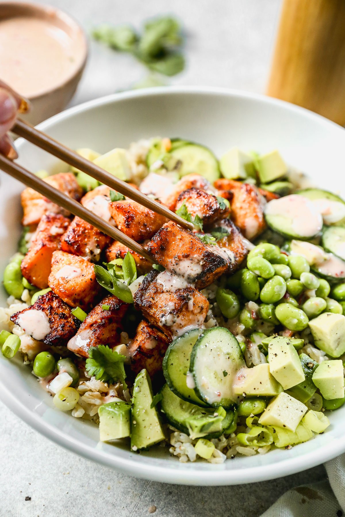 A bowl of bang bang salmon, avocado, edamame, cucumber, and rice, with chopsticks picking up a salmon piece.