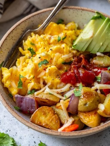 A breakfast bowl of scrambled eggs, roasted potatoes, avocado slices, cheese, salsa, and herbs, with a fork on the side.