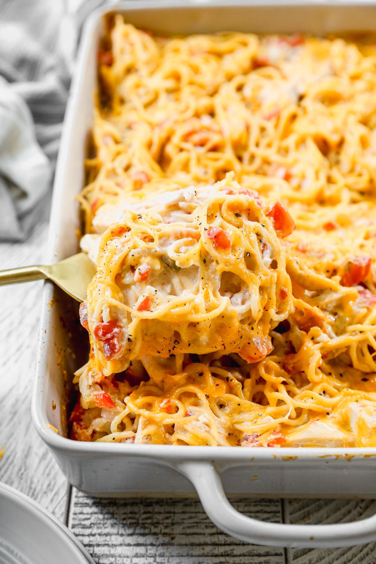 A spatula serves cheesy baked chicken spaghetti with diced tomatoes from a white casserole dish.