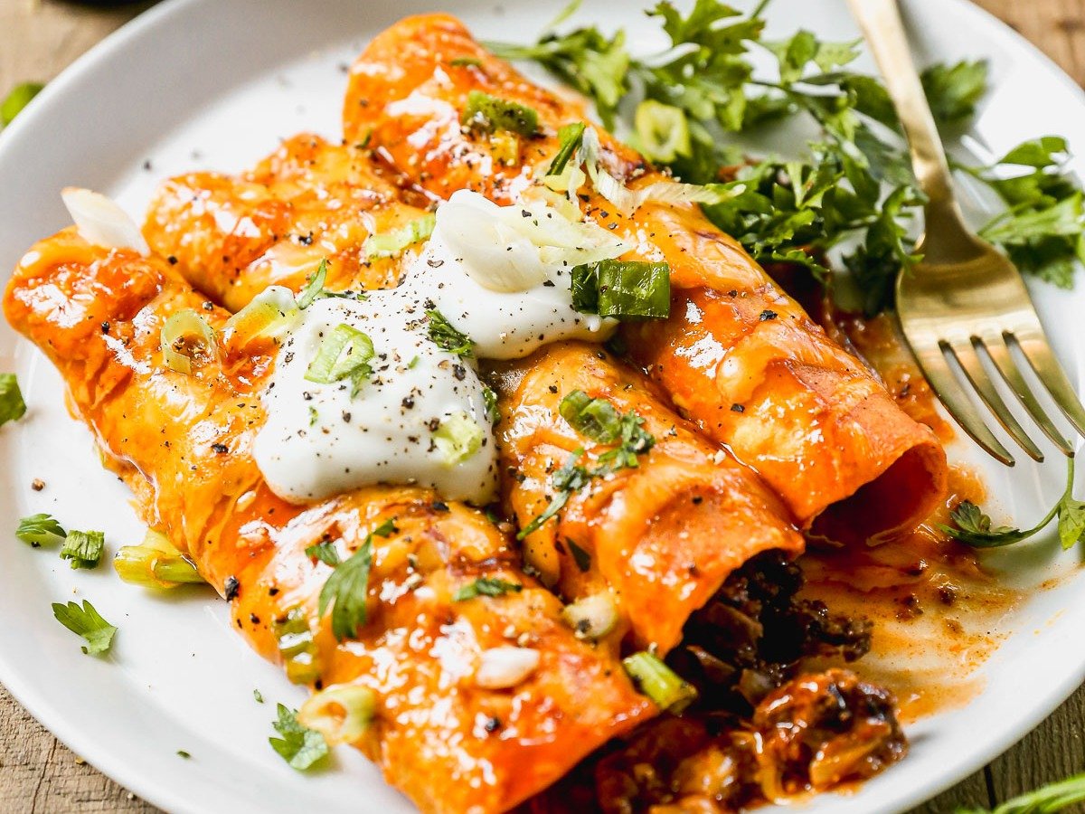 Three cheesy ground beef enchiladas topped with sour cream, green onions, and cilantro served on a white plate with a fork.