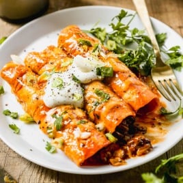 Three cheesy ground beef enchiladas topped with sour cream, green onions, and cilantro served on a white plate with a fork.