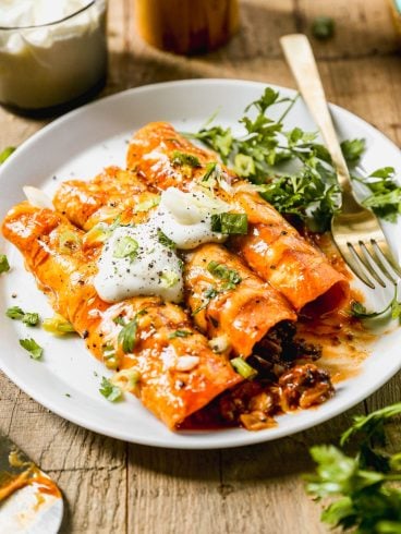 Three cheesy ground beef enchiladas topped with sour cream, green onions, and cilantro served on a white plate with a fork.