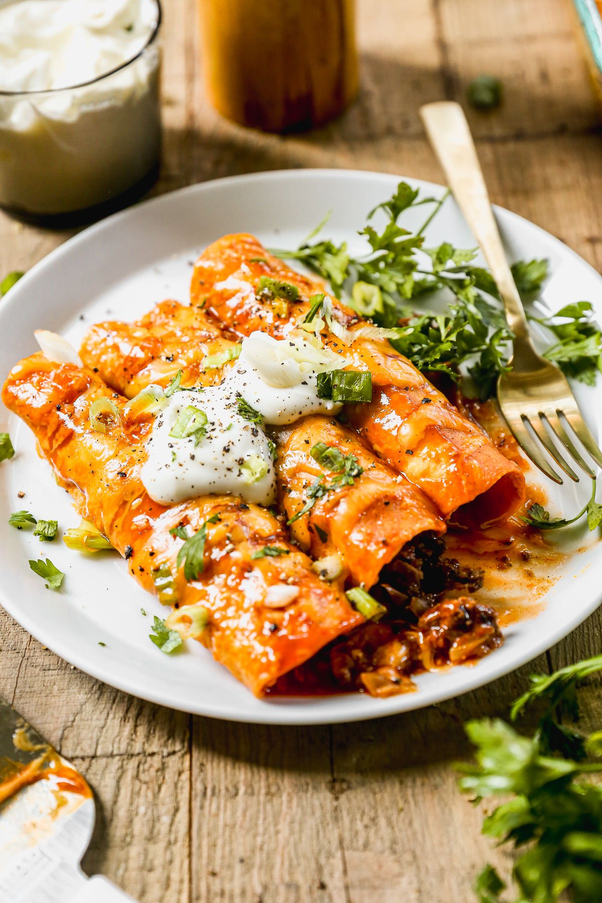 Three cheesy ground beef enchiladas topped with sour cream, green onions, and cilantro served on a white plate with a fork.