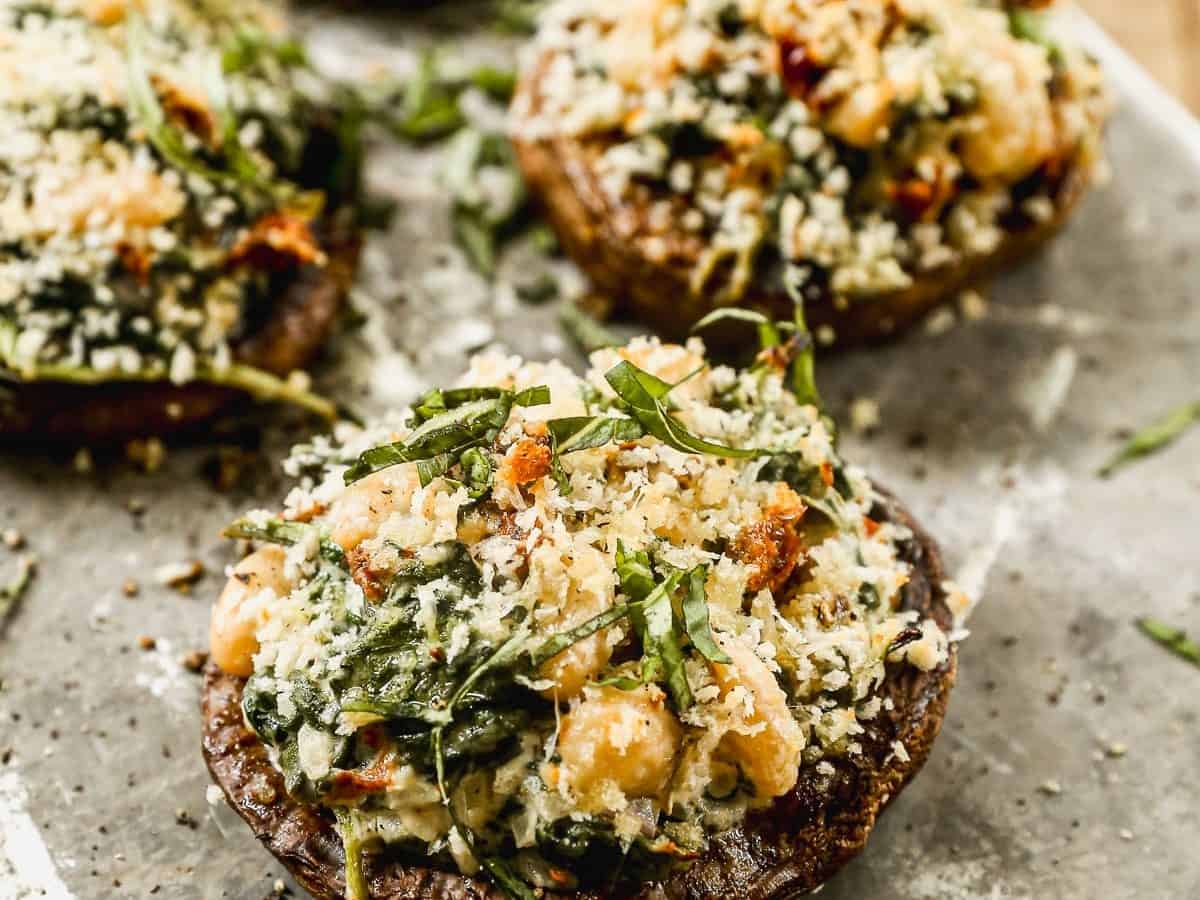Four stuffed portobello mushrooms topped with breadcrumbs and fresh herbs on a rustic metal tray.