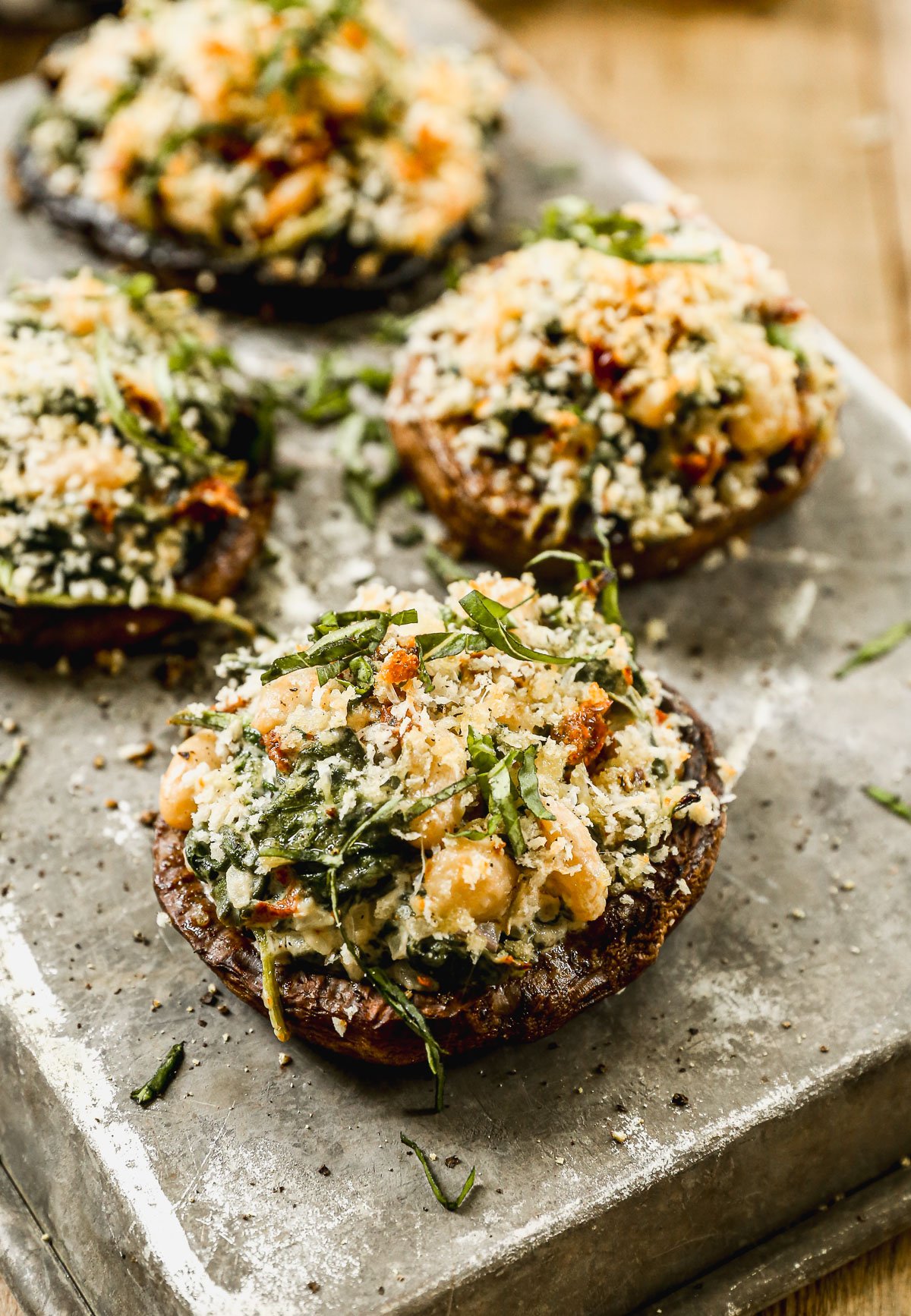 Four stuffed portobello mushrooms topped with breadcrumbs and fresh herbs on a rustic metal tray.