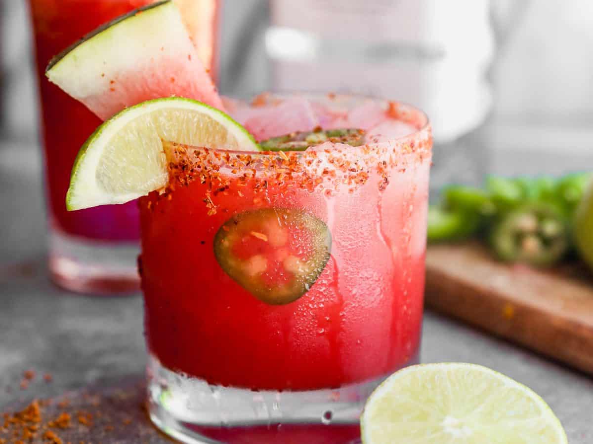 A spicy watermelon margarita with lime slices and jalapeño in a glass rimmed with chili powder, next to a halved lime.