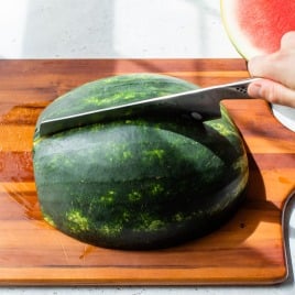 A hand slices a watermelon half on a wooden cutting board with a large knife.