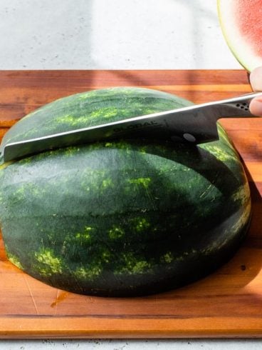 A hand slices a watermelon half on a wooden cutting board with a large knife.