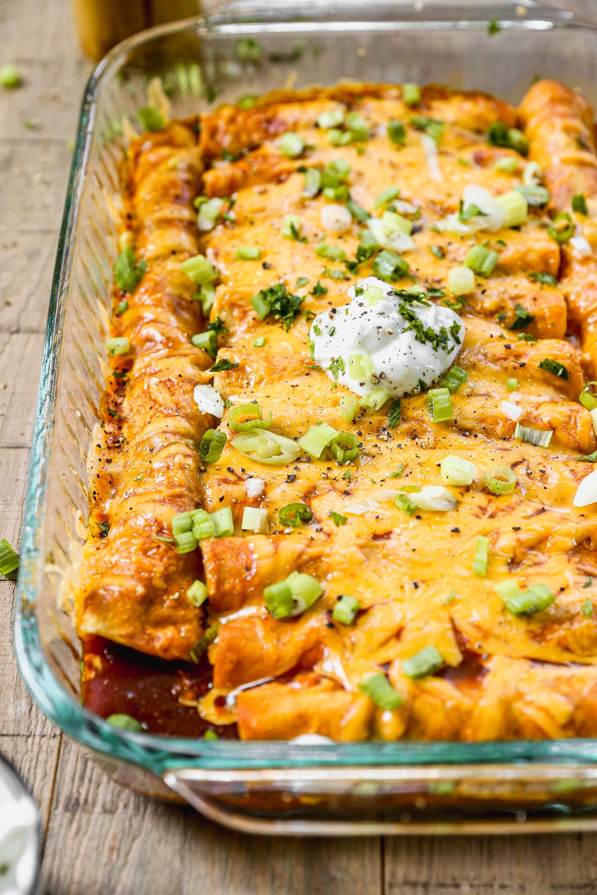 A glass baking dish of cheesy ground beef enchiladas topped with chopped green onions, cilantro, and a dollop of sour cream.