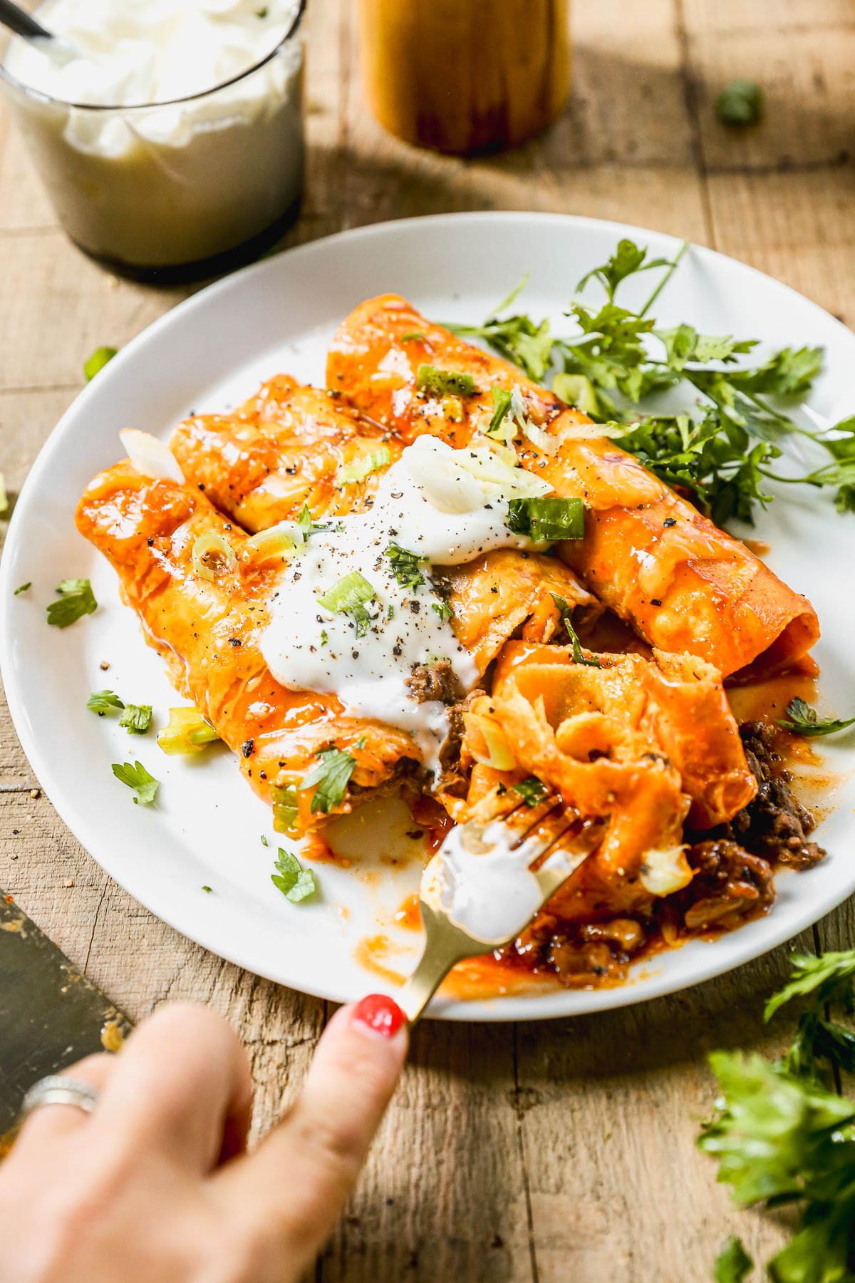 A plate of ground beef enchiladas topped with sour cream and cilantro, with a hand holding a fork taking a bite.