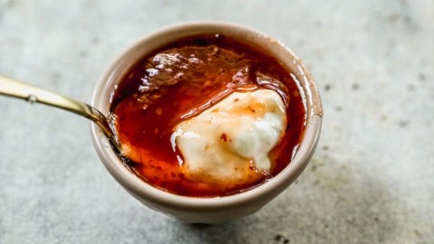 Small bowl of yogurt topped with sriracha and sweet chili sauce and a spoon, on a light textured surface.