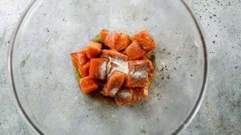 Cubed raw salmon with seasoning in a glass bowl on a gray textured surface.