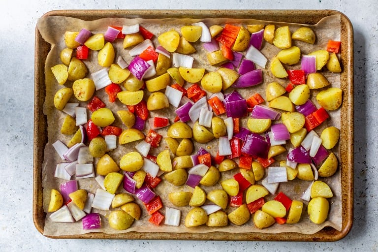 Chopped potatoes, red bell peppers, and red onions on a parchment-lined baking sheet, ready to roast.