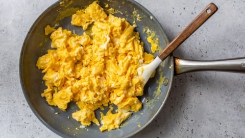 Scrambled eggs in a frying pan with a spatula on a light-colored surface.