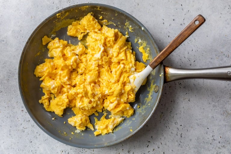 Scrambled eggs in a frying pan with a spatula on a light-colored surface.