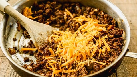 Ground beef and shredded cheddar cheese in a skillet, being stirred with a wooden spatula on a wooden surface.