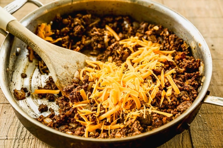 Ground beef and shredded cheddar cheese in a skillet, being stirred with a wooden spatula on a wooden surface.