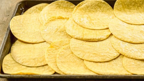 A baking sheet covered with overlapping yellow corn tortillas on a wooden surface.