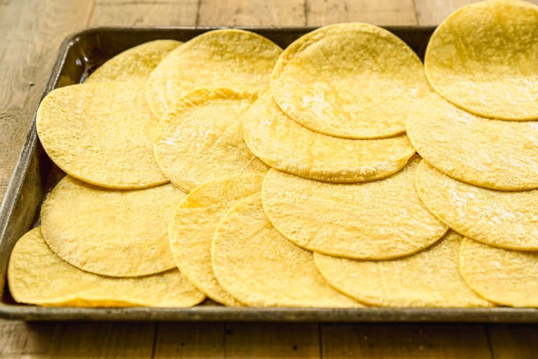 A baking sheet covered with overlapping yellow corn tortillas on a wooden surface.