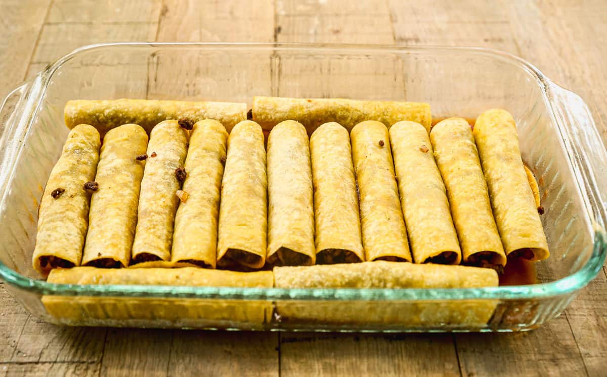 A glass baking dish filled with rolled ground beef enchiladas on a wooden surface, before adding sauce or baking.