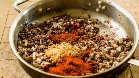 Ground beef and chopped onions cooking in a pan with piles of chili powder and other spices on top.