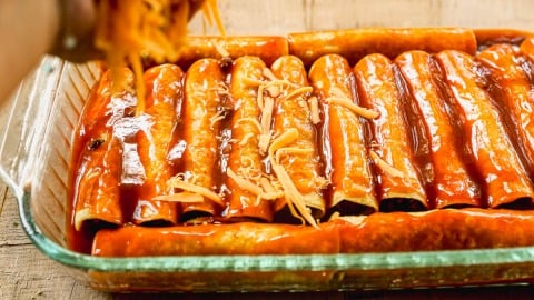 A hand sprinkles shredded cheese over rolled ground beef enchiladas covered in red sauce in a glass baking dish.