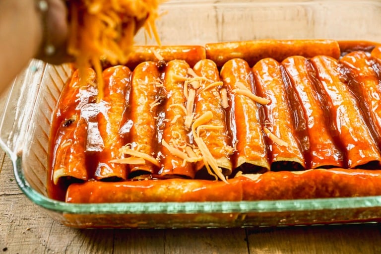 A hand sprinkles shredded cheese over rolled ground beef enchiladas covered in red sauce in a glass baking dish.